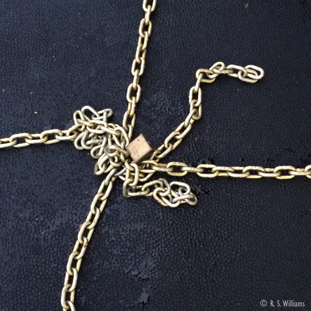 Heavy brass-plated steel chains (the kind used to secure large machinery on flatbed 18-wheeler trailers) form a cross against a black vulcanized rubber mat background. The brass-plated chains and matching lock appear neat yet messy, a sort of "bow" adorning an unexpected gift in an everyday industrial setting. 