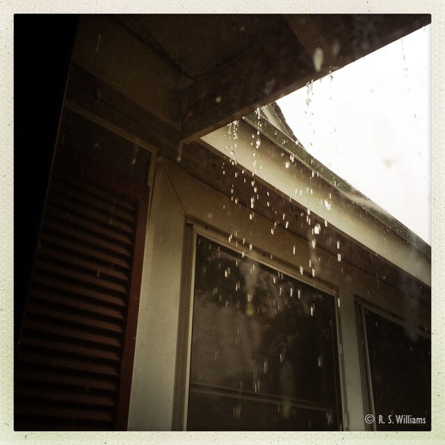 Image looking up through a glass storm door onto the porch of a house, at the corner of the roof, where two drip-edges come together. Raindrops pour off of the aluminum drip edges and down into the shot like liquid diamonds.  