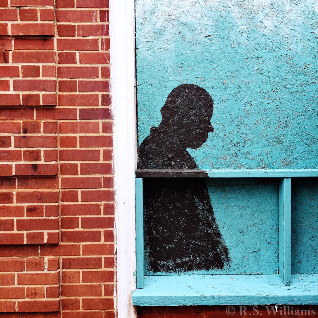 Close view of brick building facade and boarded-up corner of shop window. Plywood and 2x4 windowsill are spray-painted turquoise, with a black silhouette (in profile) of a man's head and upper torso.