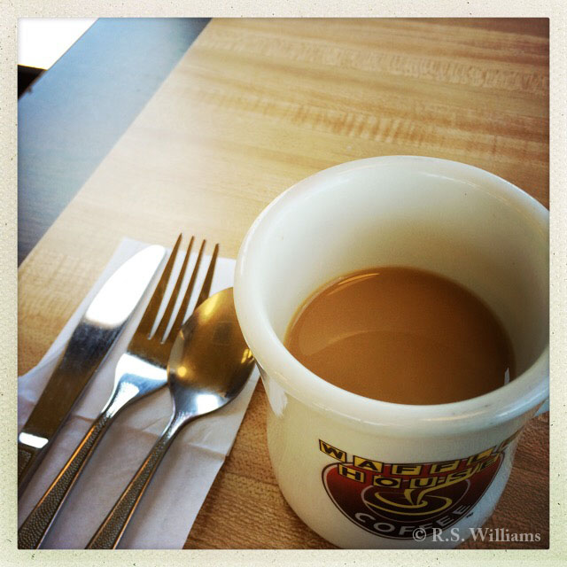 In foreground: a Waffle House logo coffee cup about half-full of light coffee, and to its left are three a knife, fork, and spoon on a white paper napkin. The rest of the frame is a wood-grain (Formica) laminate table top, light in the middle and dark around the table edges. 