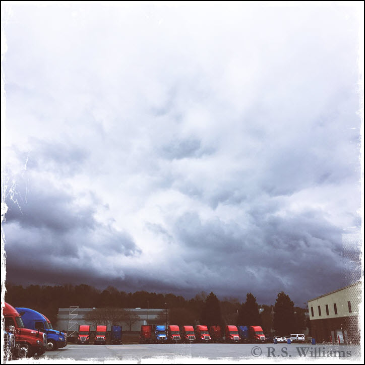 A sky filled with roiling dark gray clouds looms above a large parking area for a trucking company. In the background and at the left foreground, the dozen or so 18 wheelers (each of which is 13 feet high) and the truck garage (which stands about 30 feet high) resemble children's toys about to be swept away by the massive, looming storm behind them.
