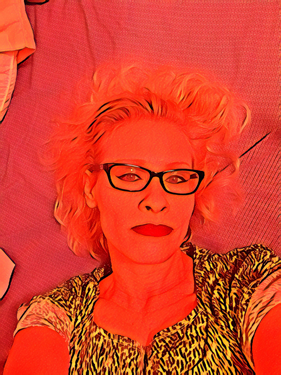 Blonde, pale-skinned woman wearing black-framed eyeglasses and tan leopard-print dress lying on patterned textiles. Other than her dress, her heavy eyeliner, and her glasses, everything in the photo is washed with bright reds and oranges, almost as if the camera lens is working with a wash of thin red paint over it.